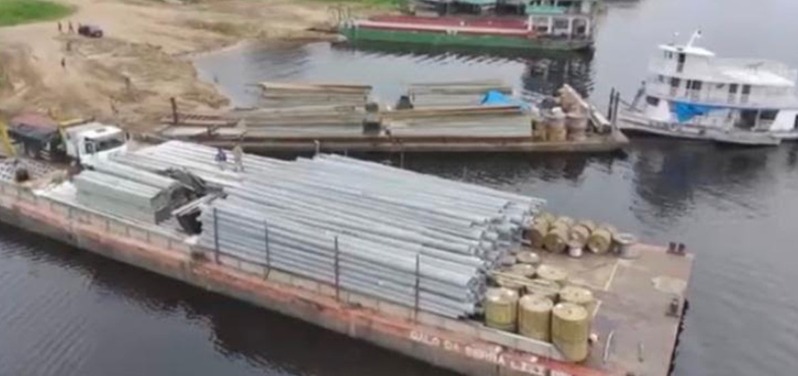 Balsa carregada com postes metálicos e bobinas de cabo em porto fluvial na Amazônia
