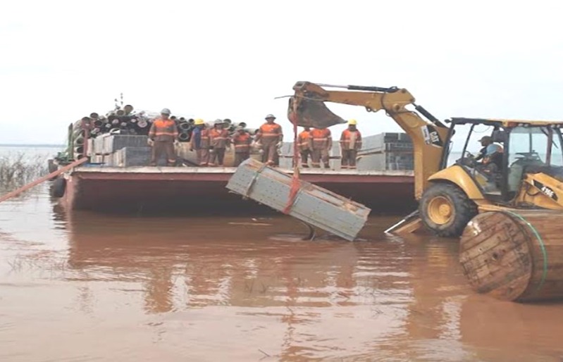 Equipe uniformizada descarregando materiais de balsa com auxílio de retroescavadeira